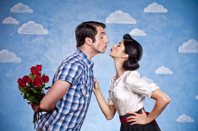 A couple coming together about to kiss. The man is holding red roses behind his back.